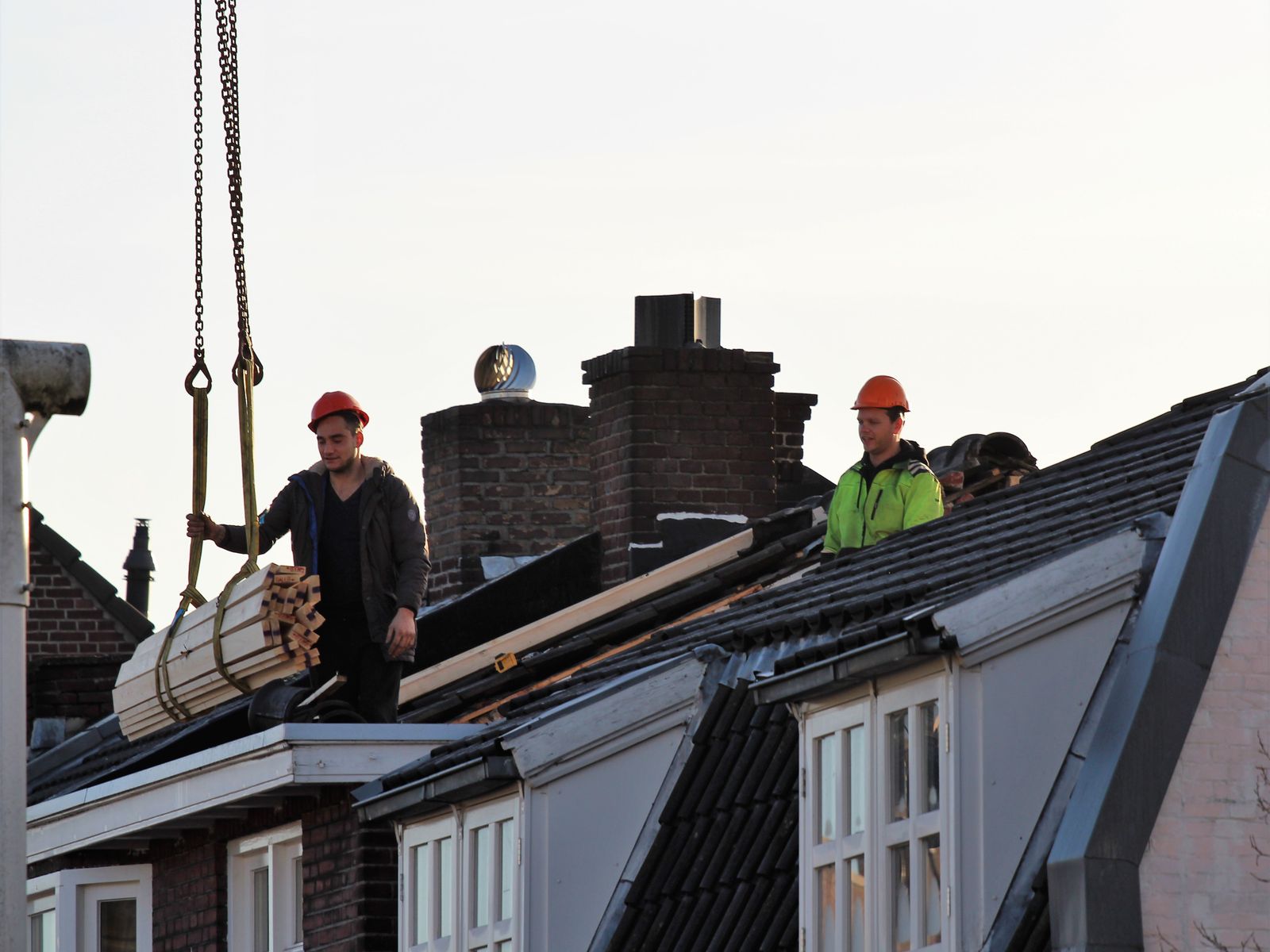 Dakdekkers aan het werk op het dak van een woning, met een kraan die materiaal aanvoert